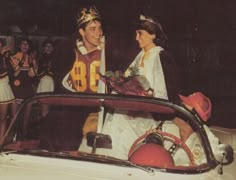 a man and woman dressed in costumes standing next to a car with other people behind them