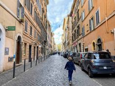 a woman walking down a cobblestone street in an old european city with cars parked on both sides