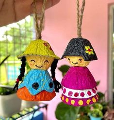 two little dolls hanging from twine strings in front of a pink wall and potted plant