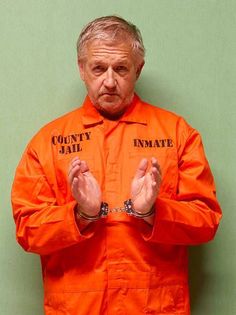 a man in an orange prison uniform with handcuffs on his hands, standing against a green wall