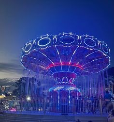 an illuminated merry go round at night with people sitting on the ground and watching it