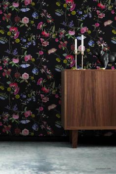 A modern interior featuring a dark floral wallpaper with colorful roses, wildflowers, and leaves, providing a vibrant backdrop. In front of the wallpaper is a wooden sideboard with a black decorative figurine and two candlesticks. A green curtain hangs to the left, and a soft rug covers the floor. Watercolour Botanical, Wallpaper Botanical, Wallpaper Textured, Illustration Agency