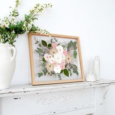 a white vase with flowers and greenery on a mantle next to a framed photograph