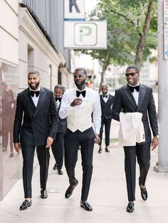 three men in tuxedos are walking down the street while looking at their cell phones