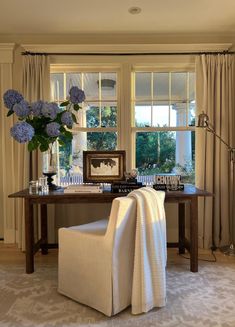 a living room filled with furniture and flowers on top of a wooden table in front of two windows