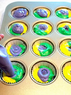 someone is decorating cupcakes with purple and green icing in a muffin tin