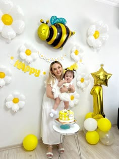 a woman holding a baby in her arms while standing next to balloons and bees on the wall