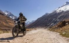 a man riding on the back of a motorcycle down a dirt road next to mountains