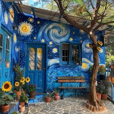 a painted house with sunflowers and potted plants on the front porch, next to a tree