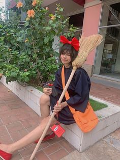 a woman sitting on the ground holding a broom and wearing a red bow, in front of a building