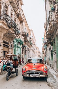 an old red car driving down a street next to tall buildings with balconies