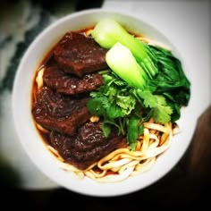 a white bowl filled with beef and noodles on top of a wooden table next to a green leafy garnish