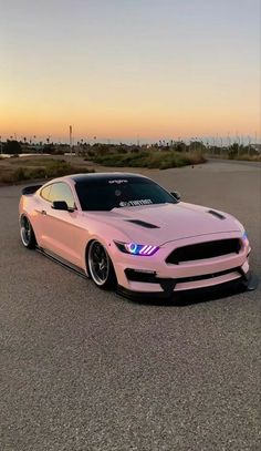 a pink mustang parked in a parking lot with the sun setting behind it and clouds