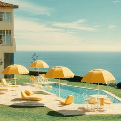 an outdoor swimming pool with yellow chairs and umbrellas next to the ocean on a sunny day