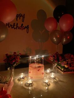 a birthday cake sitting on top of a table surrounded by candles and flowers with balloons in the background