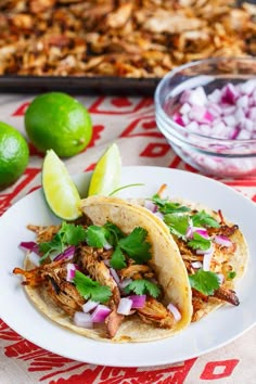 two tacos on a plate with limes, onions and cilantro in the background