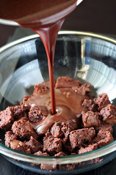 chocolate fudge being poured into a glass bowl filled with brownie squares and caramel