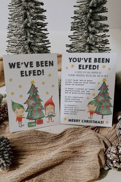 two christmas cards sitting on top of a wooden table next to pine cones and fir trees