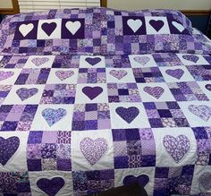 a purple and white quilt with hearts on the front, sitting on a bed in a room