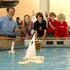 a group of kids and adults standing around a model sailboat in a water tank