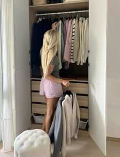 a woman standing in front of a closet with clothes on hangers and an ottoman