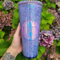 a person holding up a purple starbucks cup in front of some bushes and flowers with tattoos on their arms