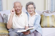 an older man and woman sitting on a couch with books in their hands, both looking at the camera