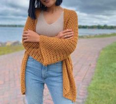 a woman standing in front of a body of water wearing a yellow sweater and jeans