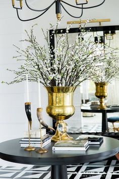 a black table topped with a gold vase filled with white flowers and greenery next to a mirror