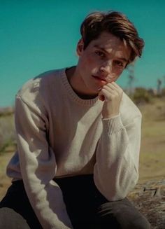 a young man sitting on top of a tree log wearing a sweater and pants with his hand under his chin