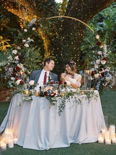 a man and woman sitting at a table with candles in front of them, surrounded by greenery