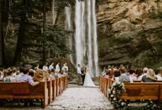 a bride and groom are standing in front of a waterfall at their wedding ceremony, surrounded by wooden pews