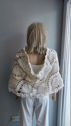 a woman standing next to a window wearing a crocheted shawl and white pants