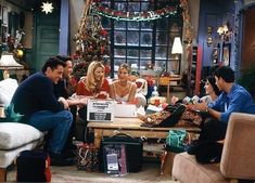 people sitting around a table in a living room with christmas decorations on the wall behind them