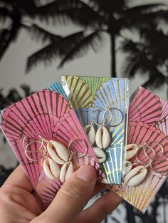 a hand holding four small cards with seashells on them and palm trees in the background