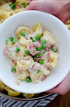 a person holding a white bowl filled with pasta, peas and ham in cream sauce