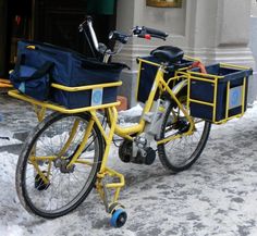 a yellow bike with two baskets on the front is parked in the snow near a building