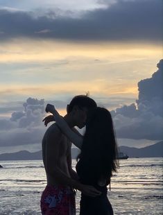 a man and woman kissing on the beach at sunset with clouds in the sky behind them