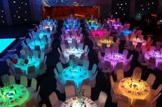 a room filled with lots of tables and chairs covered in white tablecloths, lit up by colorful lights