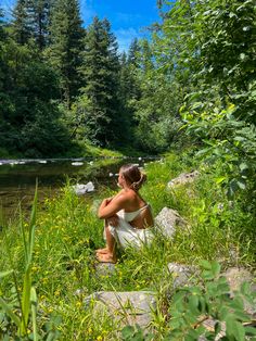 a woman in white dress sitting on rocks next to a river and looking at the water