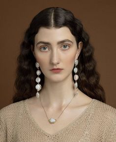 a woman with long dark hair wearing earrings and a beige sweater is standing in front of a brown background