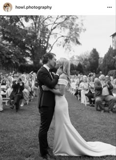 a bride and groom kissing in front of an audience at their outdoor wedding ceremony, black and white photograph