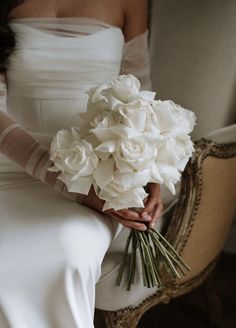 a woman in a white dress holding a bouquet of flowers sitting on a chair with her arm wrapped around the bride's waist