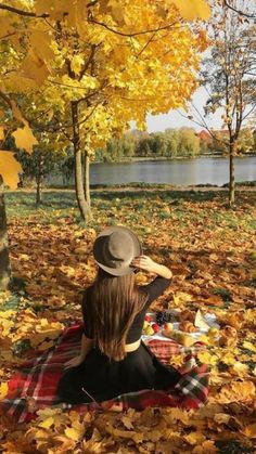 a woman sitting on top of a plaid blanket in the leaves next to a lake