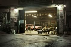 two men sitting at a table in front of an old car garage with the door open