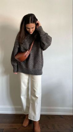 a woman standing in front of a white wall holding a brown purse and looking at the camera