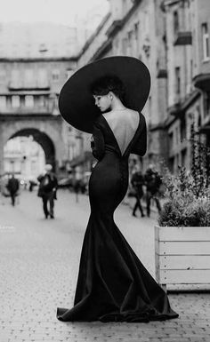 a woman in a long black dress and large hat standing on the side of a street