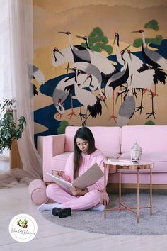 a woman sitting on the floor reading a book in front of a wall with cranes painted on it