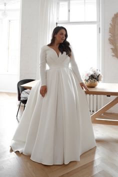 a woman standing in front of a window wearing a white wedding dress with long sleeves