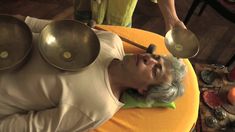 an older woman is sitting on a yellow chair with two large metal pots over her head